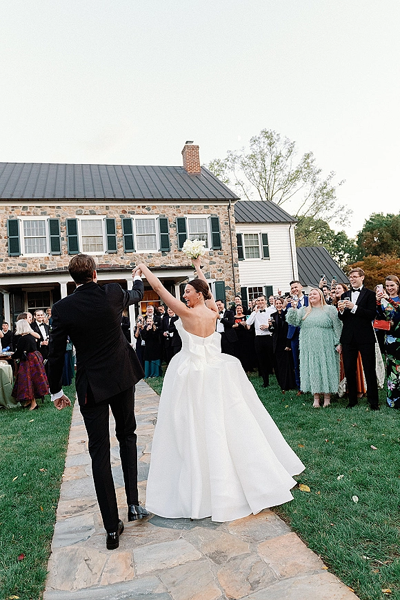 Wedding recessional as bride and groom walk away hand in hand, bride lifts bouquet on stone path while guests cheer by estate house