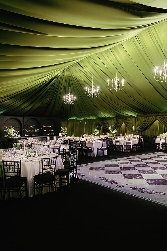 Reception decor under a draped tent ceiling with chandeliers over round tables in white linens, black chiavari chairs, candles, and floral centerpieces