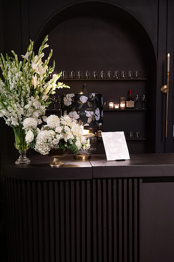 Wedding welcome table with a wedding welcome sign display, white florals in glass and brass vases, candlelight, and lamp before black bar shelves