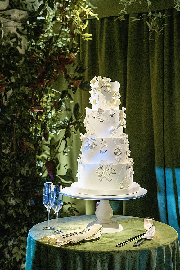 Wedding cake with white buttercream floral applique on a white stand atop a green velvet table, champagne flutes and candles before green drapery