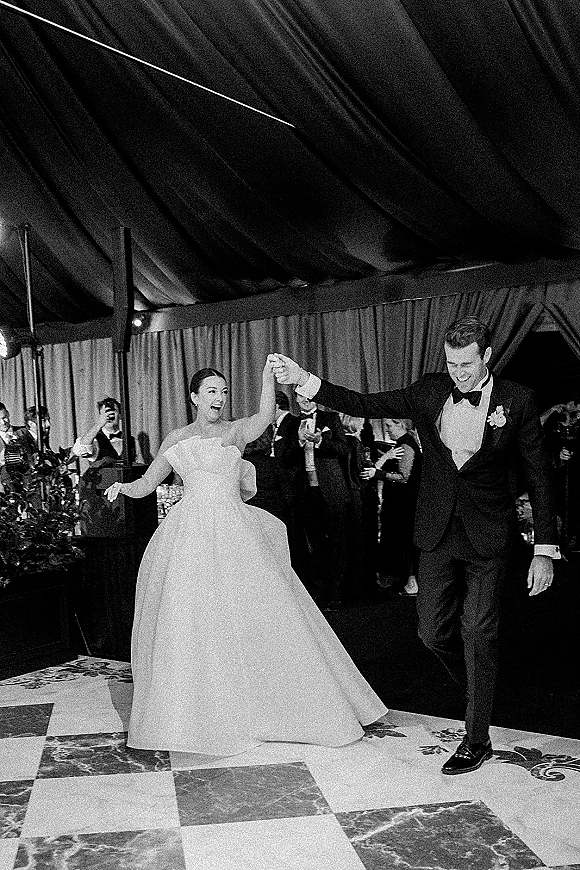 Reception entrance as newlyweds hold hands, raising arms on a checkered dance floor under a draped tent ceiling with cheering guests