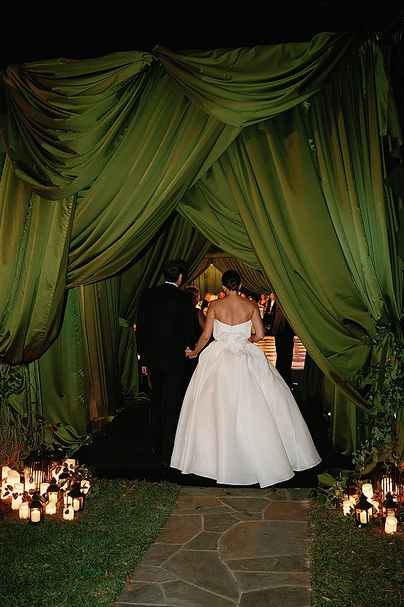 Couple entrance as newlyweds walk hand in hand into a draped tent, candlelit walkway lined with lanterns and greenery behind them
