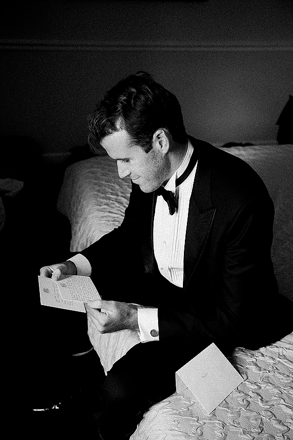 Groom portrait in a black tuxedo reading a handwritten letter on a bed in a bedroom, holding an envelope in a quiet moment