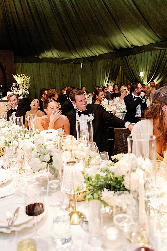 Wedding reception guests reacting to a toast, laughing at candlelit tables in a draped tent with white floral centerpieces and greenery garlands
