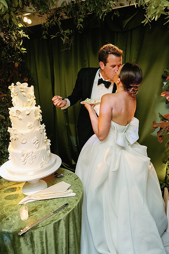 Cake cutting moment with bride in strapless bow dress and groom in tuxedo beside tiered cake on green velvet table under foliage arch