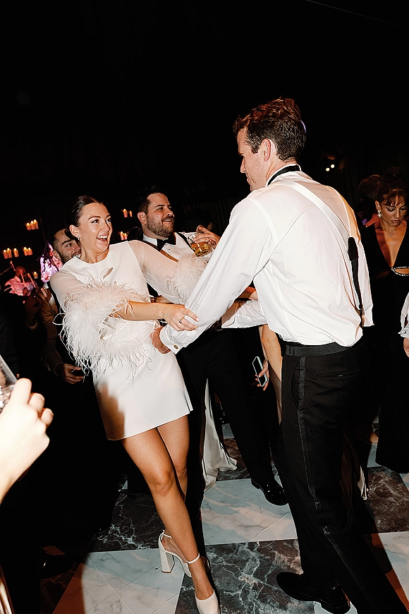 Wedding reception dancing as bride in a white mini wedding dress with feather cuffs dances with groom in suspenders on a candlelit floor