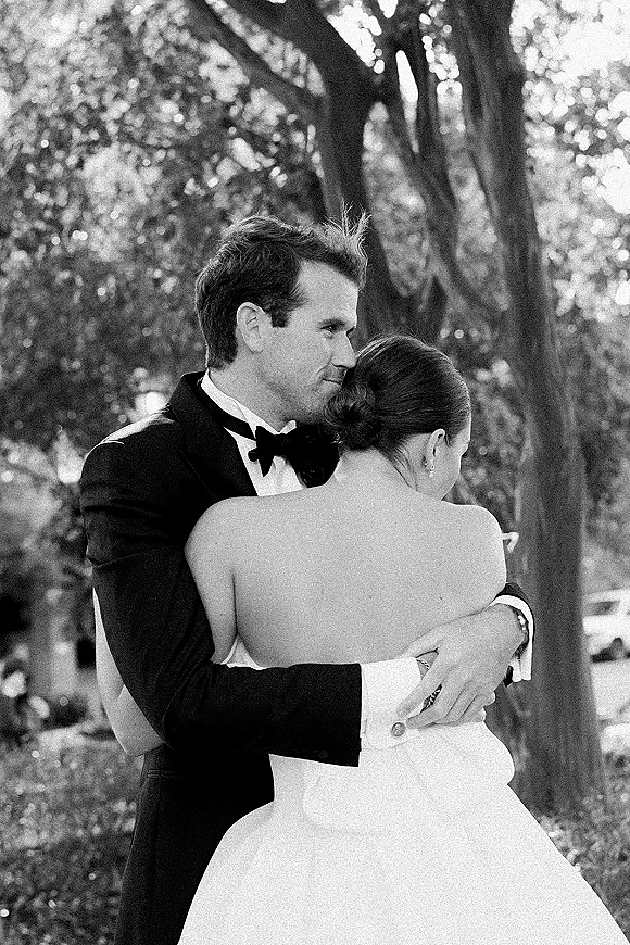 Couple portrait of bride and groom embrace, her strapless wedding dress and pearl earrings against a tree-lined park backdrop