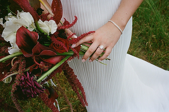 Bridal bouquet with burgundy calla lilies, red roses and cascading amaranthus in bride’s hands, ring and bracelet on a lawn