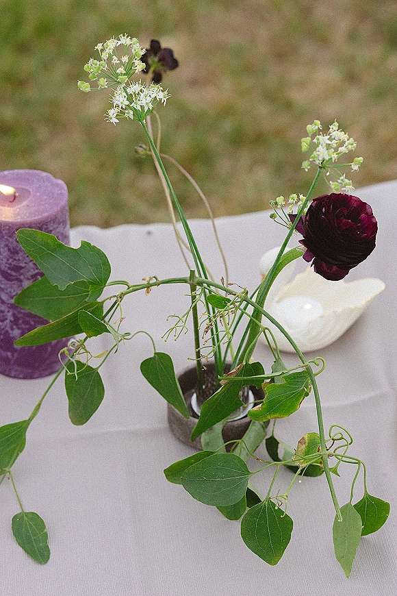 Wedding centerpiece with a bud vase of white wildflowers and greenery vines, accented by a purple pillar candle on linen in a grassy field