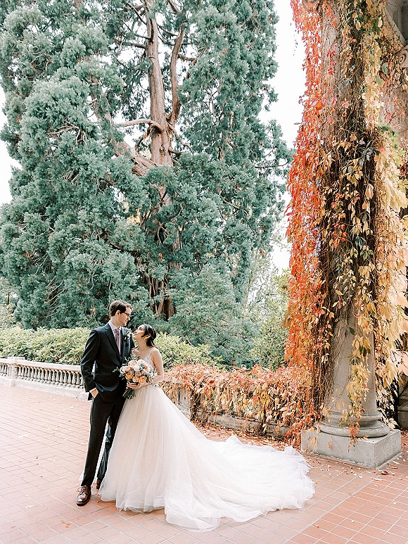 Couple portrait of bride and groom outdoors, bride holding a pastel bouquet and gazing up at him by ivy stone columns in autumn