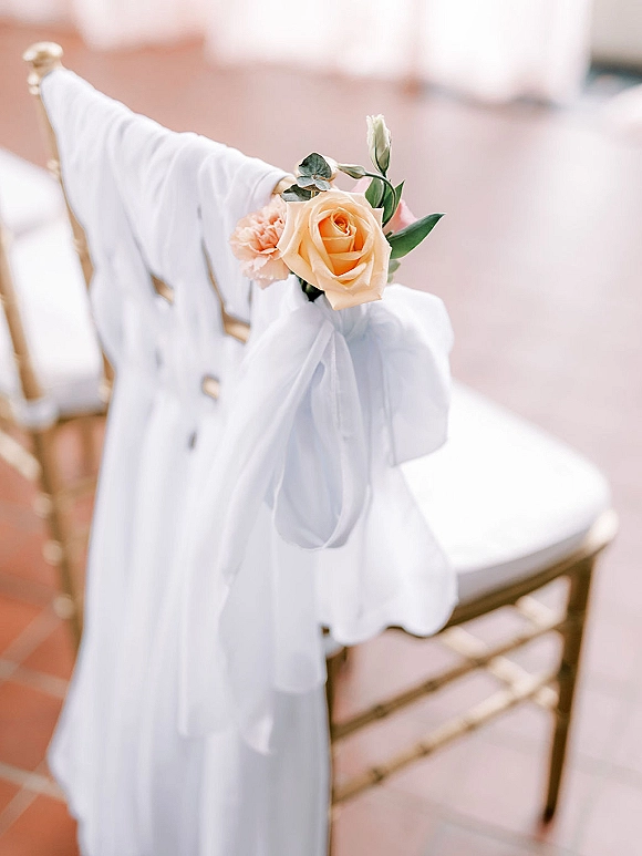 Ceremony chair decor with white chiffon chair drape, tied with a peach rose, blush carnation, and greenery on gold chairs over terracotta tile floor