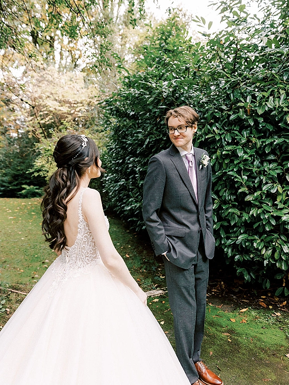 First look moment as bride in lace wedding dress approaches groom in suit with glasses, holding hands in a garden with hedges and trees