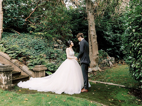 Couple portrait of bride and groom holding hands, bride looking up at him, her long train and veil on garden steps by ivy wall