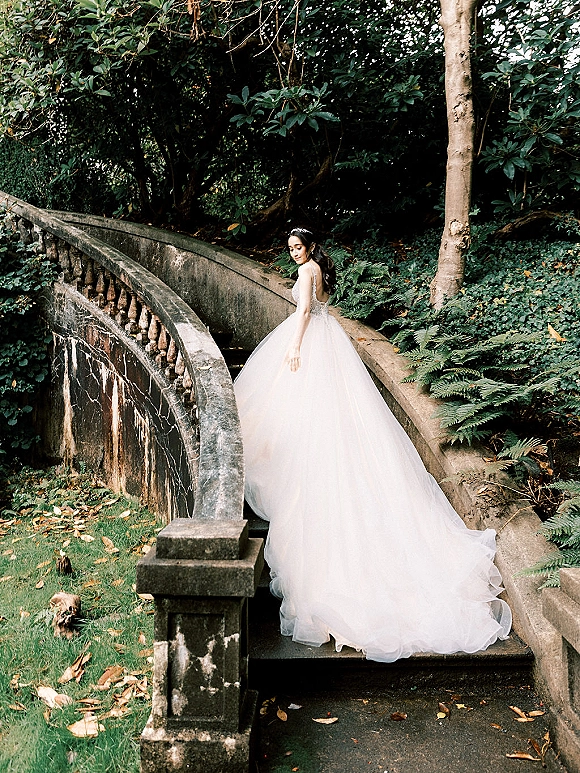 Bridal portrait of a bride in a strapless lace gown with long train, looking back on stone bridge steps amid lush garden greenery and trees