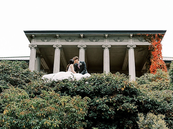 Wedding kiss portrait of bride and groom kissing in tuxedo and flowing gown train on steps before ivy-covered columns and garden shrubs