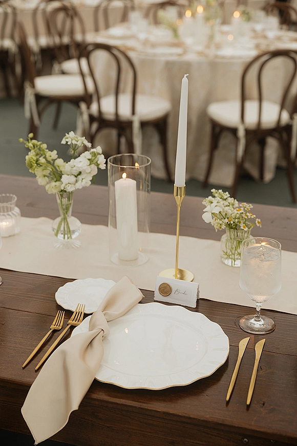Reception tablescape with wedding table setting featuring white taper candles, bud vases, scalloped plates, gold flatware, and wax-seal place cards on round tables with bentwood chairs