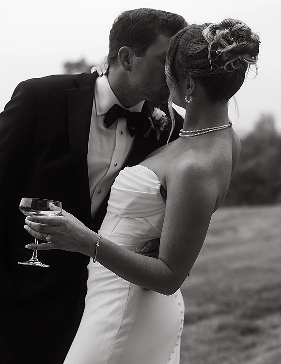 Wedding kiss portrait of bride and groom kissing, bride in strapless dress with pearls holding a champagne coupe in a field with trees
