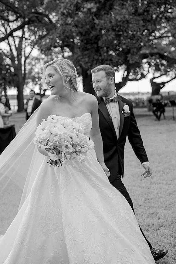Newlywed couple portrait of bride and groom walking hand in hand, bride holding bouquet with veil flowing on a tree-lined lawn