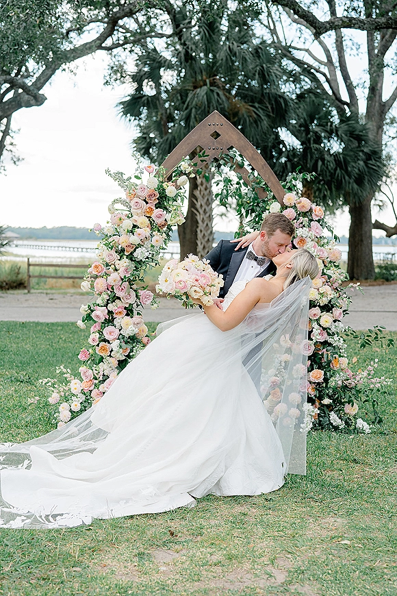 Wedding kiss portrait of bride and groom kissing in a romantic dip under a blush rose floral arch by the waterfront, her long veil flowing