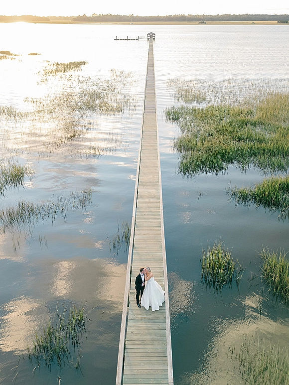 Wedding kiss portrait of bride and groom kissing on a wooden dock over a lake at sunset, her dress train flowing behind