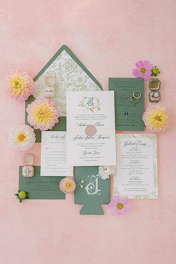 Invitation suite wedding invitation flat lay with sage green cards, wax seal, rings, earrings, and floral blooms on a pink backdrop