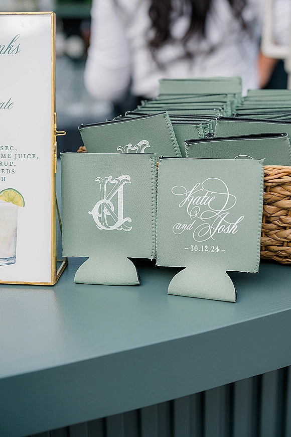 Wedding koozies with custom wedding koozies calligraphy names and date in a wicker basket beside a gold-framed drink menu at the bar counter