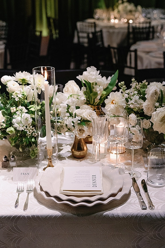 Reception tablescape with wedding table centerpiece of white florals, taper candles in glass hurricanes, scalloped plates and menu cards in a dim room