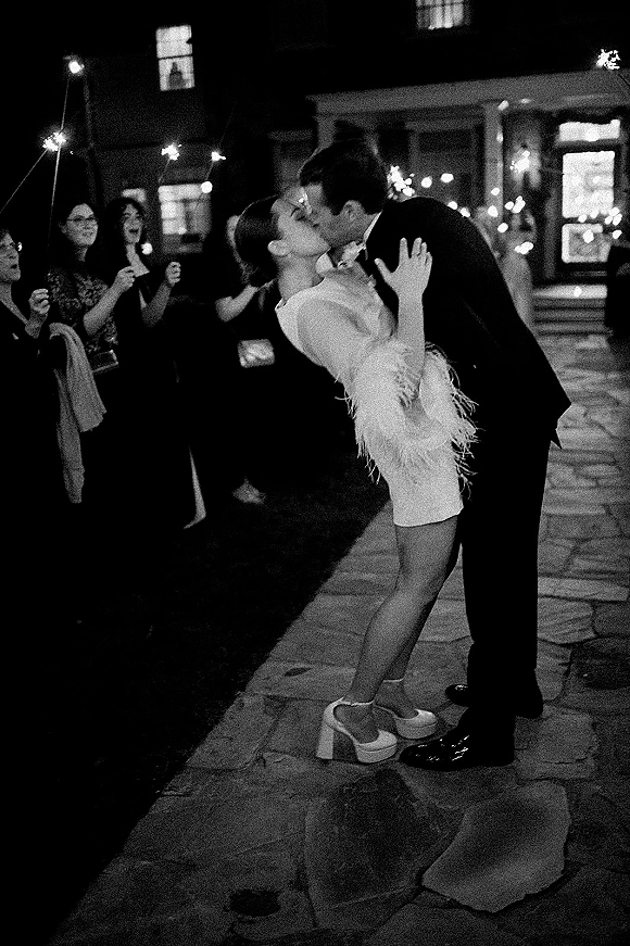 Wedding kiss portrait during a sparkler exit as the groom dips the bride in a feather-trim mini dress on a stone patio under string lights at night