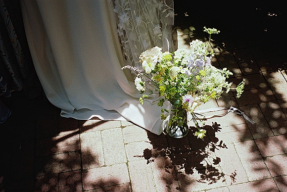 Bridal bouquet of white and green wildflowers in a glass vase with ribbon beside a lace wedding dress hem on sunlit stone tiles