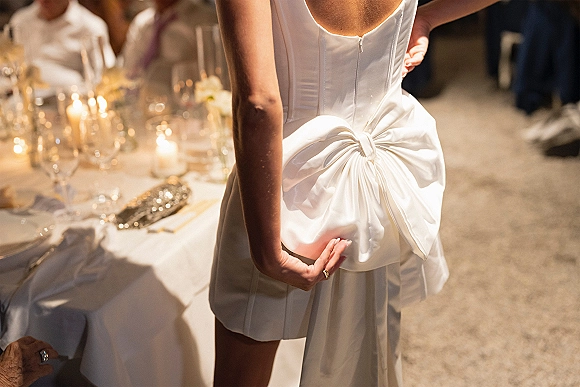 Wedding dress back in a strapless satin wedding dress with an oversized bow and ring detail beside candlelit reception table glassware
