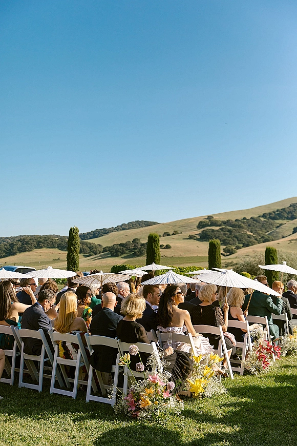 Wedding ceremony guests seated outdoors with white parasols, sunglasses, and floral aisle arrangements on a lawn with rolling hills beyond