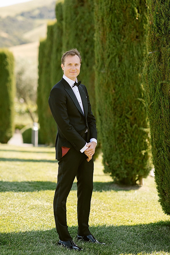 Groom portrait in a classic groom tuxedo, standing full length with hands clasped on a lawn beside cypress trees and hills