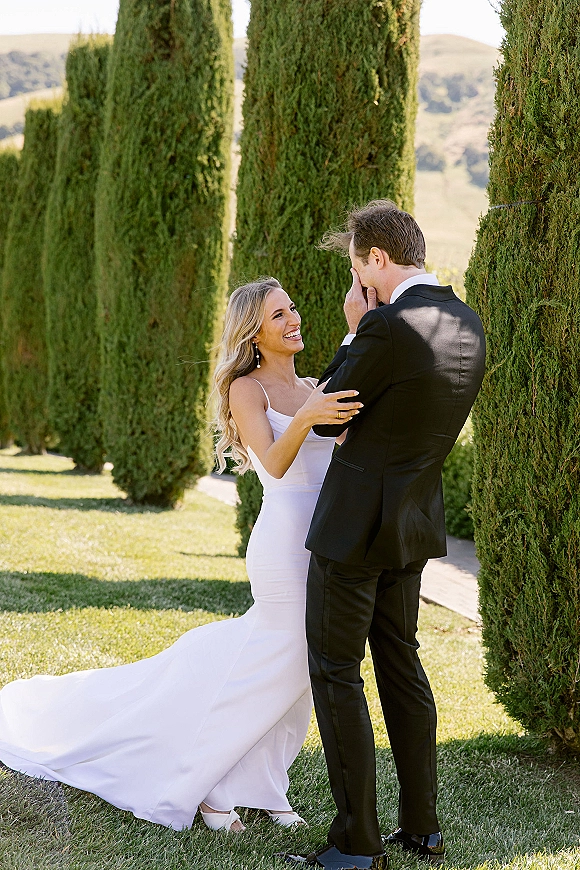 Wedding first look as bride touches groom’s face, his emotional reaction beside cypress-lined garden path, her dress train flowing