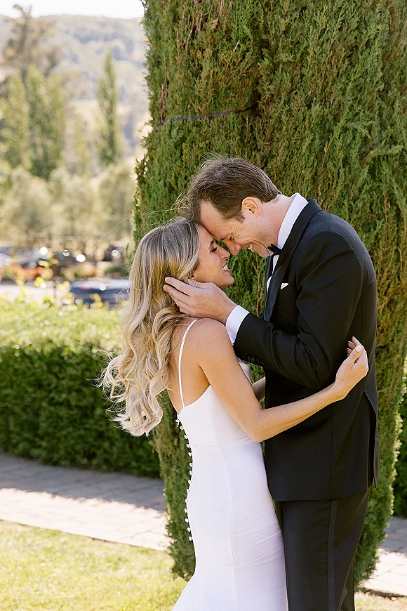 Couple portrait of bride and groom embrace, foreheads touching as he holds her face on a garden path, cypress trees behind