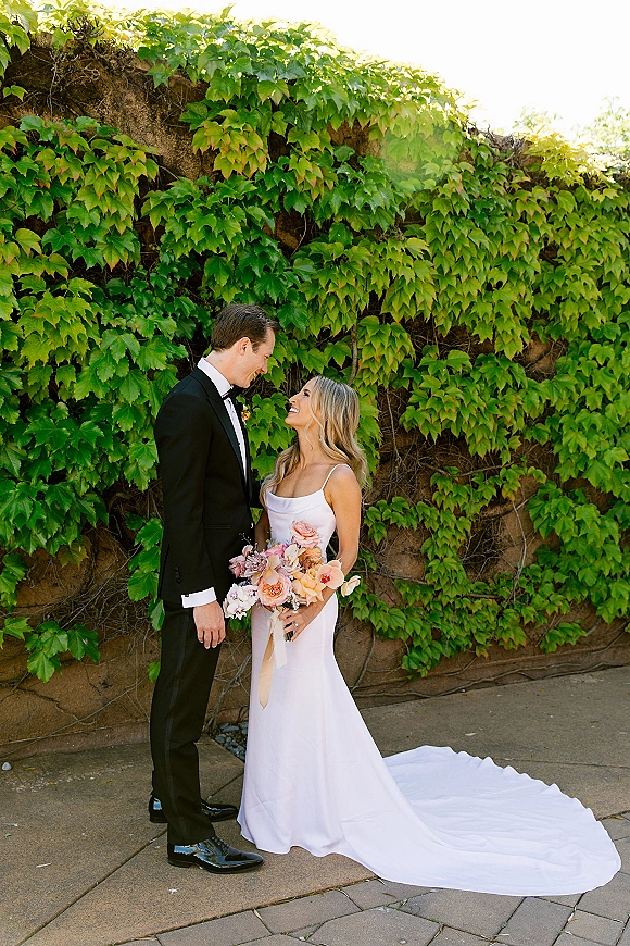 Couple portrait of bride holding bouquet, smiling up at groom in black tuxedo beside an ivy-covered stone wall walkway