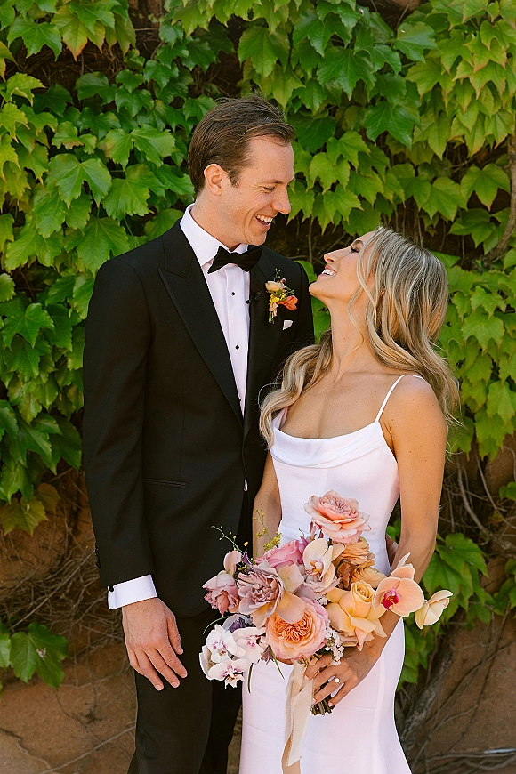 Couple portrait of bride holding a blush and peach orchid bouquet, smiling up at groom in black tuxedo before an ivy stone wall