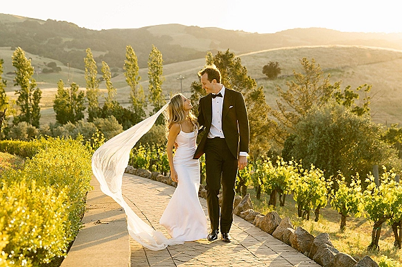 Couple portrait of bride and groom walking on a stone path as her wedding veil blows, backlit by golden sunlight in a vineyard hills setting