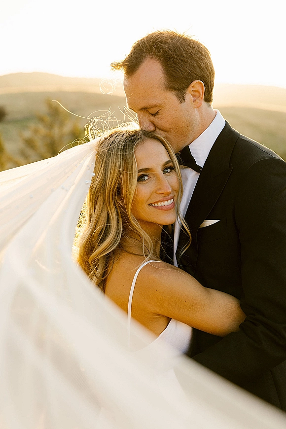 Couple portrait of bride and groom embrace as he kisses her forehead, long veil blowing in wind against sunset mountains backdrop