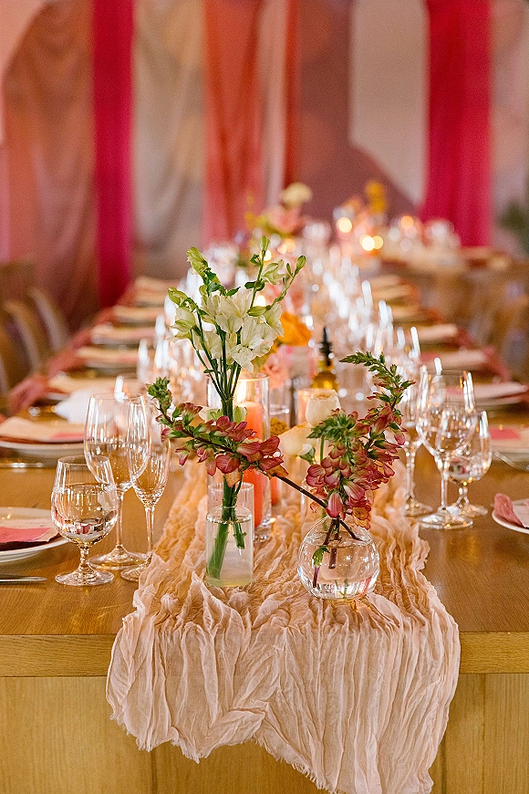 Reception tablescape on a long banquet table setting with wildflower bud vases, taper candles, gauze runner, and draped fabric backdrop