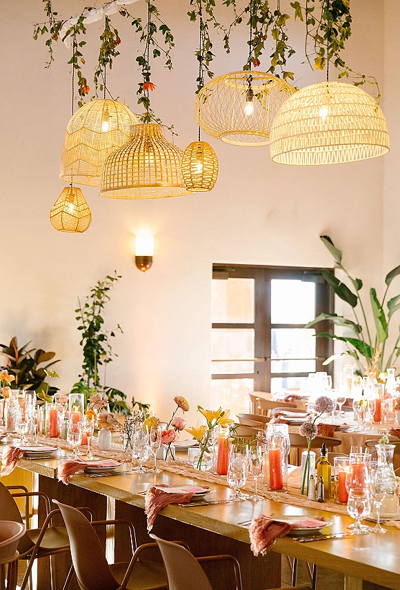 Reception tablescape on a long wedding table with bud vases, floral centerpieces, pink taper candles, and hanging greenery under wicker pendants