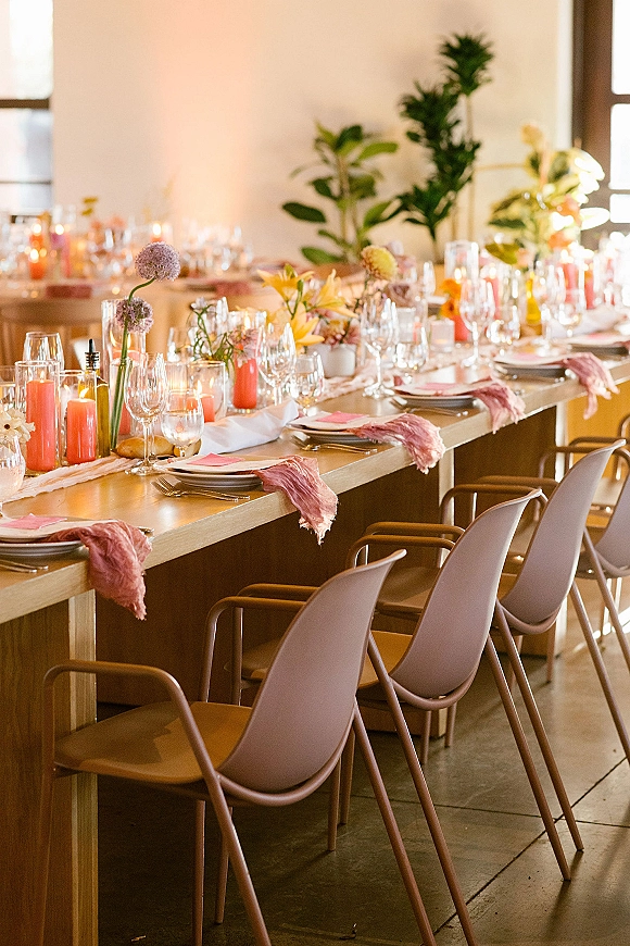 Reception tablescape on a long banquet table wedding with pink taper candles, floral bud vases, and place settings by large windows
