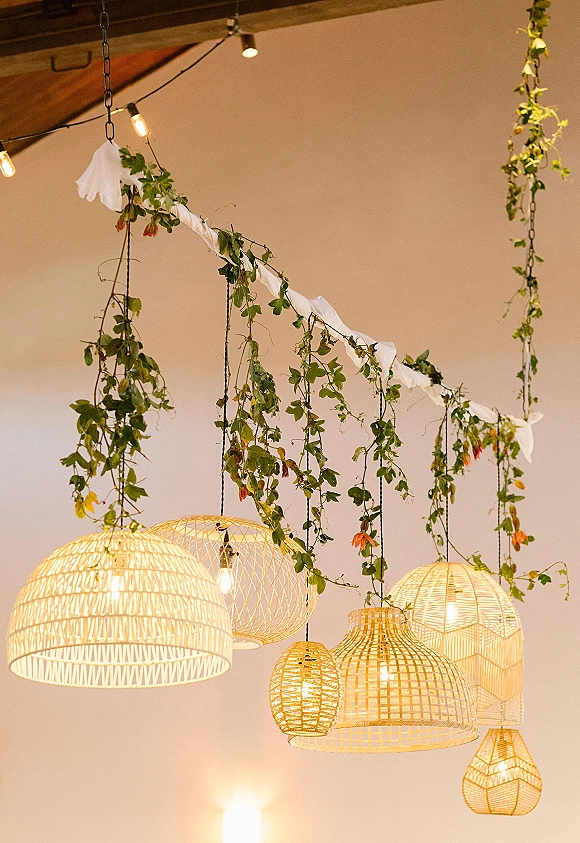 Hanging wedding lighting with rattan pendant lamps and draped fabric, paired with greenery garlands and string lights under wood beams