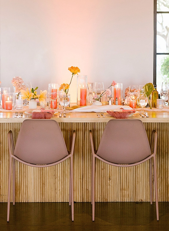Reception tablescape with wedding head table decor featuring colorful taper candles, bud vases, and mixed glassware on a wood table by a window