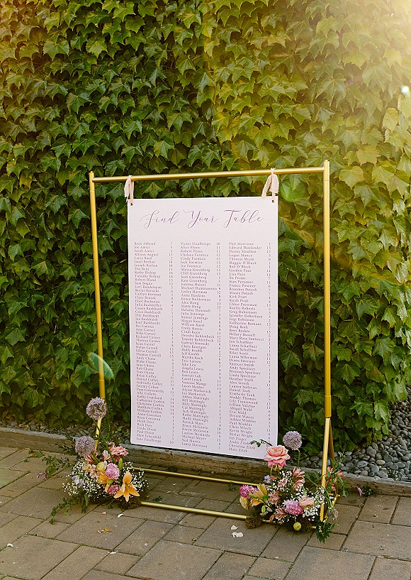 Wedding seating chart on a gold seating chart stand with ribbon ties and florals, set against an ivy wall with stone pavers and river rocks