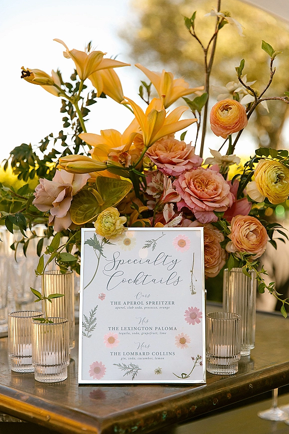 Wedding bar signage with a specialty cocktails sign beside pastel florals in glass vases on a sunlit outdoor bar cart among trees