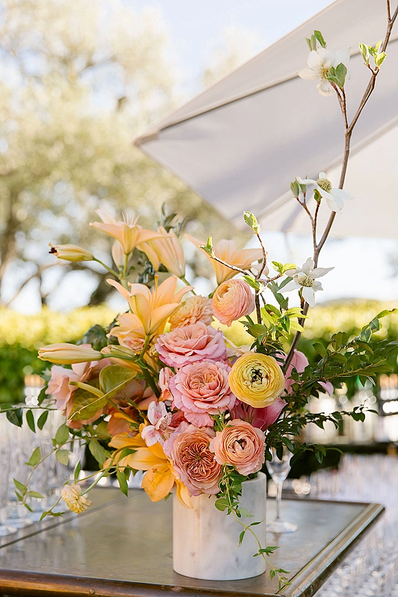 Wedding centerpiece flowers in a white vase with peach lilies and pink roses on a mirrored table, dogwood branches, garden patio backdrop