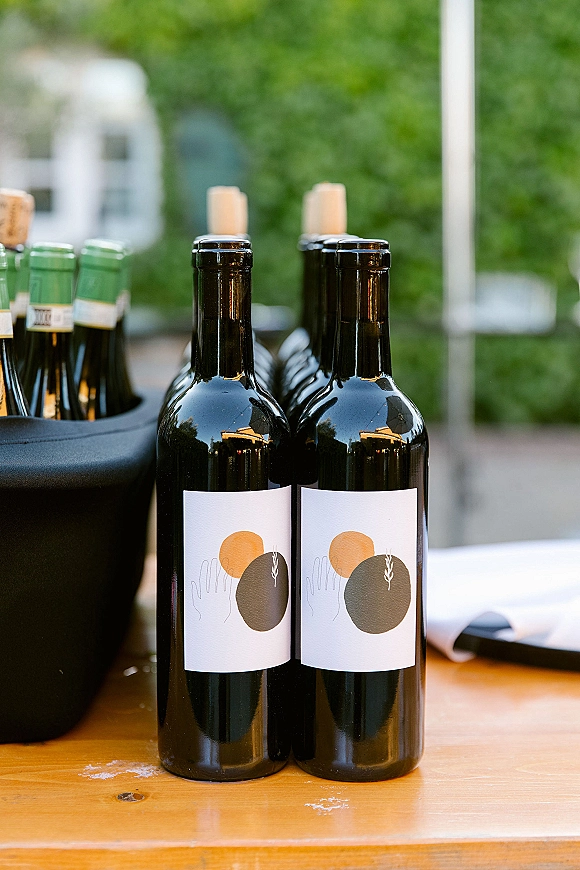Wine bottles at a wedding drink station on a wooden table, with custom labels, corks, opener, and chiller tub on an outdoor patio