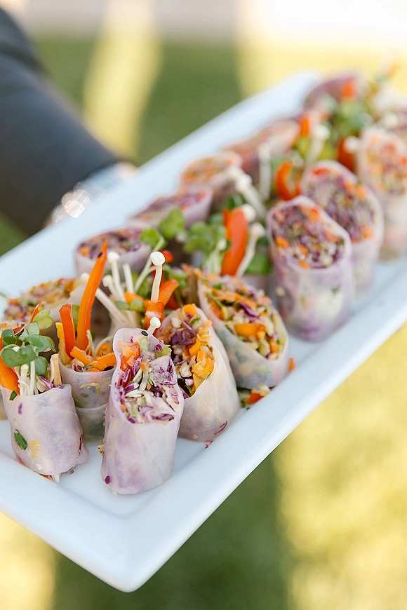 Wedding appetizers arranged on a white serving platter with spring rolls, microgreens, shredded carrots and purple cabbage on a blurred lawn background