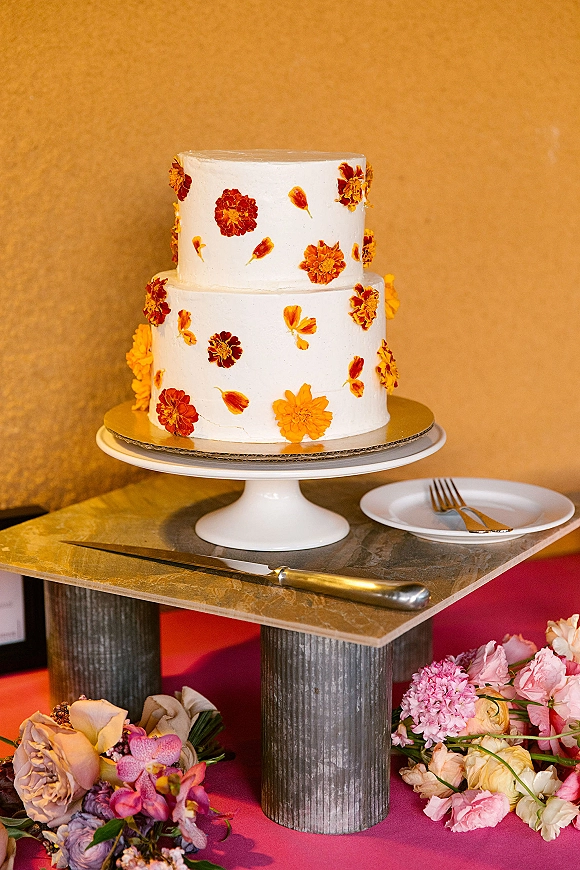 Wedding cake with white buttercream on a pedestal stand, decorated with marigold petals and edible flowers, beside knife and plates on tabletop