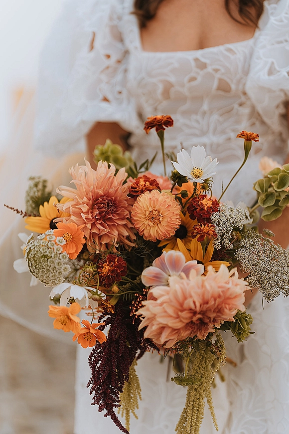 Bridal bouquet with colorful wildflower bouquet of dahlias, marigolds, cosmos and greenery held against lace bodice and veil on neutral backdrop